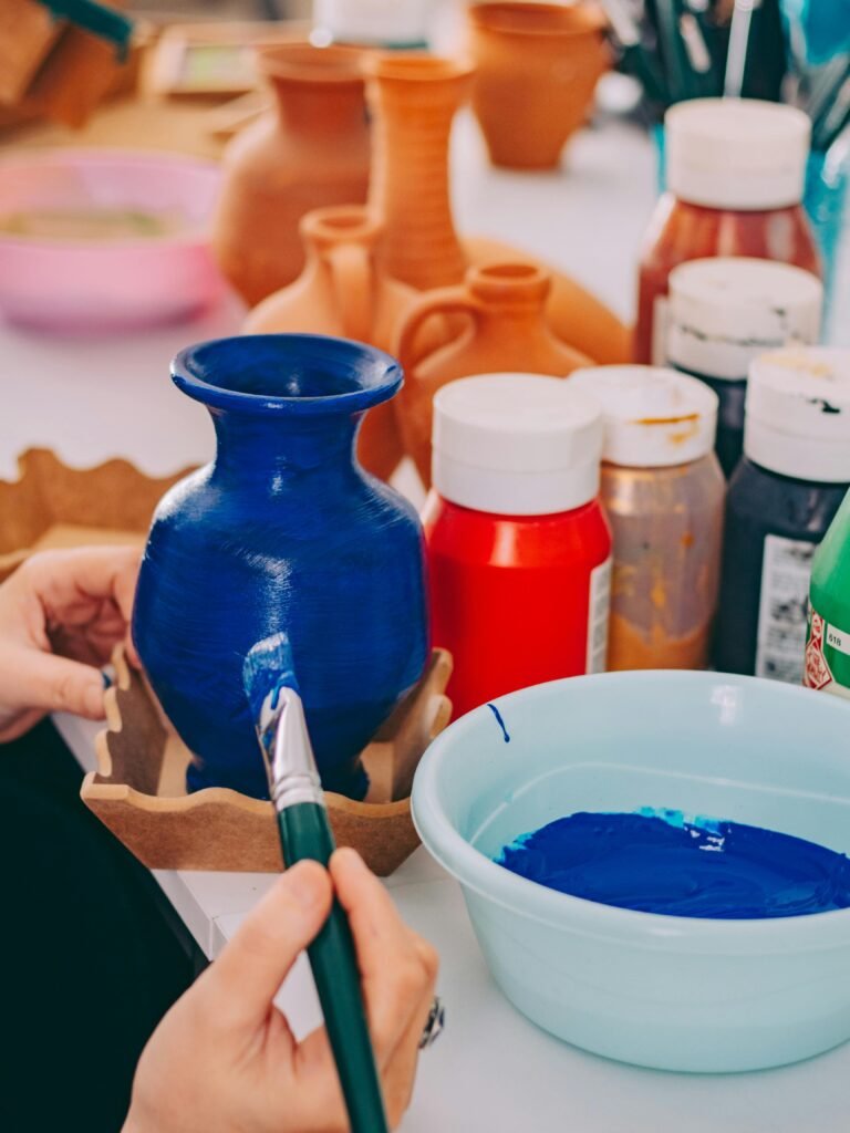 A person carefully paints a ceramic vase blue surrounded by vibrant paints and pottery.