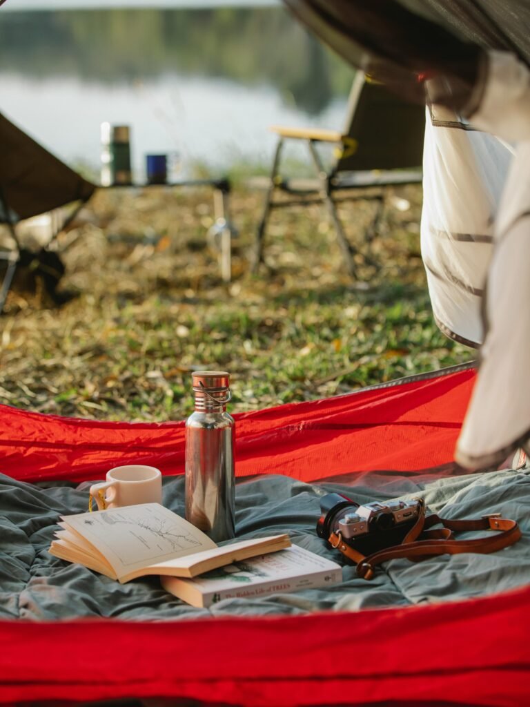 Camp tent with book thermos and photo camera arranged on grassy peaceful lakeside on clear summer day